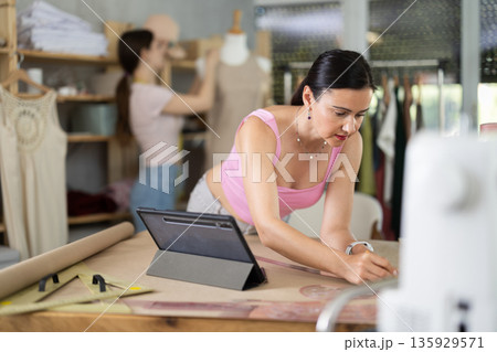 Female tailor drawing clothing outlines on paper in sewing studio Female tailor drawing clothing outlines on paper in sewing studio 135929571