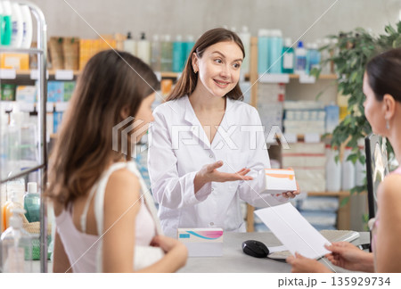 Attentive young female pharmacist advising family of mother and daughter choosing box with pills in pharmacy store Attentive young female pharmacist advising family of mother and daughter choosing box with pills in pharmacy store 135929734