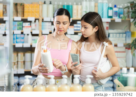 Portrait of attentive mother and daughter customers holding and selecting cream or skin care product in drugstore Portrait of attentive mother and daughter customers holding and selecting cream or skin care product in drugstore 135929742