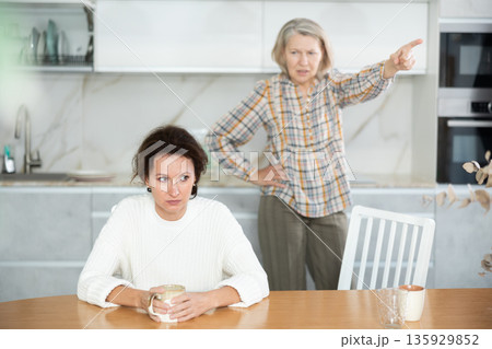 Upset middle-aged woman sitting at the kitchen table with her back to old woman quarreling to her 135929852