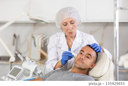 Female cosmetologist performing anti-aging face phototherapy procedure for male client in clinic of aesthetic clinic Female cosmetologist performing anti-aging face phototherapy procedure for male client in clinic of aesthetic clinic 135929855