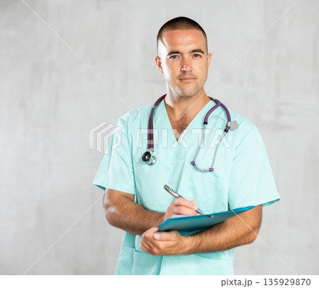Portrait of male doctor in medical gown with stethoscope making notes in patient medical history Portrait of male doctor in medical gown with stethoscope making notes in patient medical history 135929870