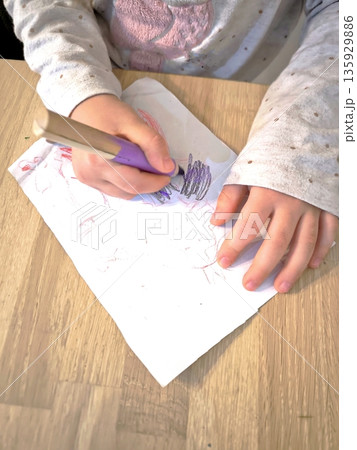 A child draws on white paper with a wax crayon, viewed from above. 135929886