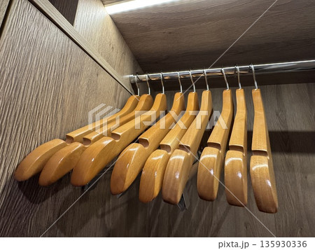 Wooden clothes hangers hanging on a metal rod inside a wooden wardrobe, with an LED light strip above. Wooden clothes hangers hanging on a metal rod inside a wooden wardrobe, with an LED light strip above. 135930336