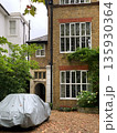 London brick townhouse facade with large white windows, ivy, arched entry, gravel yard and a car covered with a grey tarp. 135930364