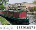 Green and red narrowboat moored on a calm canal, ropes and fenders visible, with brick apartment building and balconies behind. 135930373