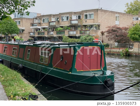 Green and red narrowboat moored on a calm canal, ropes and fenders visible, with brick apartment building and balconies behind. 135930373