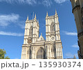 London view of Westminster Abbey west towers with clock, Gothic stone facade and a flagpole against a clear blue sky with clouds. 135930374