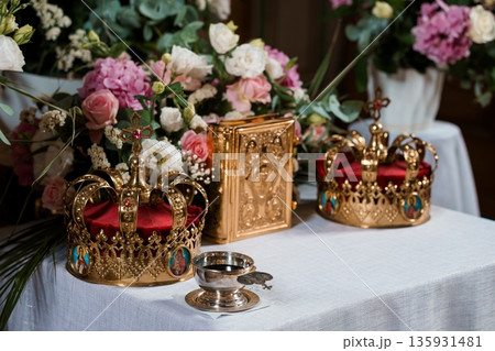 Golden Wedding Crowns and Religious Icons on White Table with Floral Arrangement in Church Setting 135931481