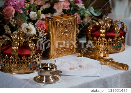 Golden Orthodox wedding crowns, ornate religious icons, floral arrangements, and ceremonial items on white cloth with soft lighting Golden Orthodox wedding crowns, ornate religious icons, floral arrangements, and ceremonial items on white cloth with soft lighting 135931485