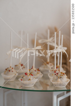 Elegant White Cake Pops with Gold and Red Sprinkles on Glass Table with Delicate Ribbon Ties Elegant White Cake Pops with Gold and Red Sprinkles on Glass Table with Delicate Ribbon Ties 135931500