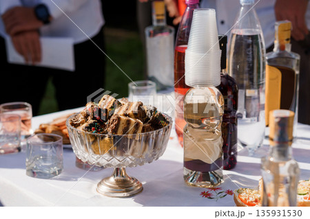 Outdoor Party Table with Assorted Desserts, Bottled Beverages, and Plastic Cups in Sunlit Setting 135931530