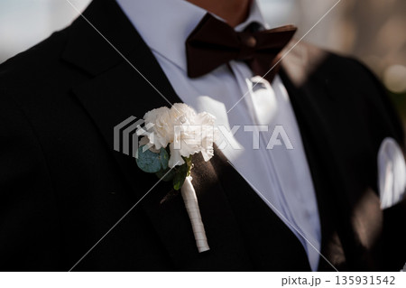 Elegant Groom Wearing Black Tuxedo with White Carnation Boutonniere and Brown Bow Tie 135931542
