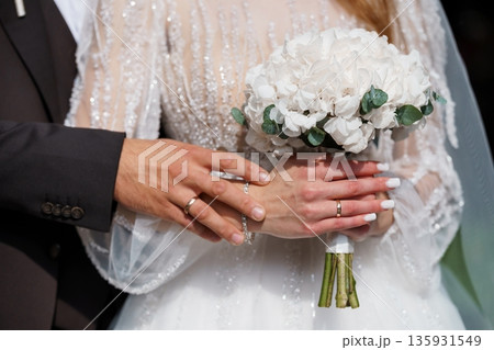 Elegant wedding couple holding hands with white hydrangea bouquet and rings in close-up view 135931549