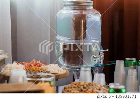 Large Glass Water Dispenser with Metal Faucet on Buffet Table Surrounded by Snacks and Drinks Large Glass Water Dispenser with Metal Faucet on Buffet Table Surrounded by Snacks and Drinks 135931571