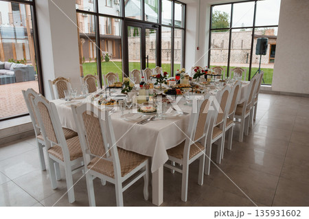 Elegant White Table Setting with Floral Arrangements in Modern Glass-Walled Dining Room with Outdoor Garden View Elegant White Table Setting with Floral Arrangements in Modern Glass-Walled Dining Room with Outdoor Garden View 135931602