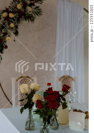 Elegant Wedding Table Setting with Red and White Roses in Glass Vases Under Floral Arch Decoration 135931605