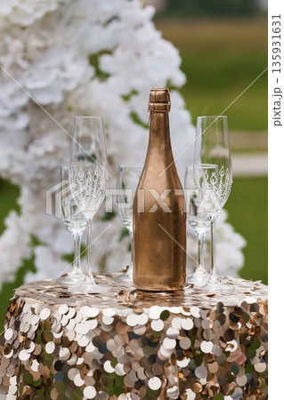 Elegant Gold Champagne Bottle and Crystal Flutes on Sequin Table with White Floral Arch Background Elegant Gold Champagne Bottle and Crystal Flutes on Sequin Table with White Floral Arch Background 135931631