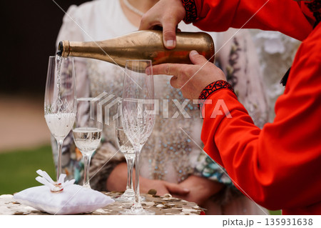 Traditional Ukrainian wedding ceremony with couple pouring golden champagne into ornate glasses amidst festive attire and confetti Traditional Ukrainian wedding ceremony with couple pouring golden champagne into ornate glasses amidst festive attire and confetti 135931638