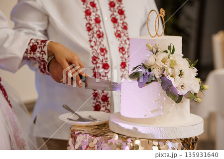 Bride and groom cutting wedding cake together in traditional Ukrainian embroidered attire at elegant celebration event Bride and groom cutting wedding cake together in traditional Ukrainian embroidered attire at elegant celebration event 135931640