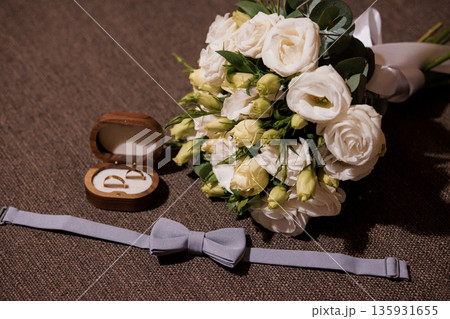 Elegant Wedding Bouquet with White Roses and Eustoma, Wooden Ring Box, and Gray Bow Tie on Brown Textured Background Elegant Wedding Bouquet with White Roses and Eustoma, Wooden Ring Box, and Gray Bow Tie on Brown Textured Background 135931655