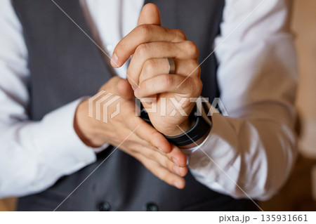 Close-up of a groom adjusting his wedding ring and wristwatch in a formal white shirt and black vest Close-up of a groom adjusting his wedding ring and wristwatch in a formal white shirt and black vest 135931661