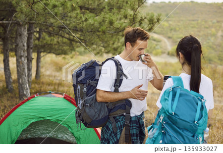Couple of tourists camping with backpacks near forest tent, man drinking from mug Couple of tourists camping with backpacks near forest tent, man drinking from mug 135933027