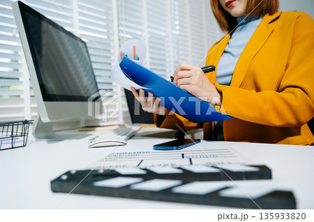Creative female filmmaker working with tablet, clapperboard, and laptop in modern studio office. Film production, planning, and content creation scene. 135933820