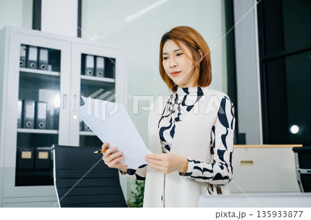 Young Asian woman in office analyzing paper reports, planning strategy with laptop and documents. Young Asian woman in office analyzing paper reports, planning strategy with laptop and documents. 135933877