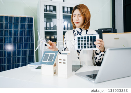 Asian businesswoman in office holding green globe with solar house and wind turbine models symbolizing renewable energy, sustainability Asian businesswoman in office holding green globe with solar house and wind turbine models symbolizing renewable energy, sustainability 135933888