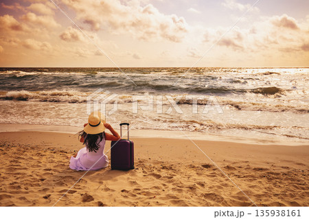 Seascape during sunrise. A woman sits on the beach in summer with a travel suitcase. The girl looks at the sea. Travel, happy holiday, summertime concept Seascape during sunrise. A woman sits on the beach in summer with a travel suitcase. The girl looks at the sea. Travel, happy holiday, summertime concept 135938161