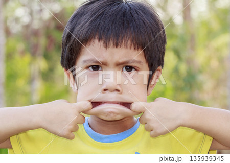 Portrait of Asian little boy doing a funny pose pulling the corner of his mouth with his fingers outdoor 135939345