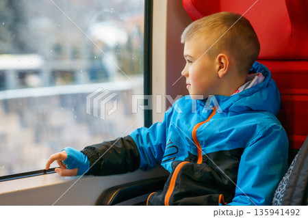 Thoughtful young boy in warm jacket sitting by train window, looking outside during winter travel. Concept of childhood, transportation, journey Thoughtful young boy in warm jacket sitting by train window, looking outside during winter travel. Concept of childhood, transportation, journey 135941492