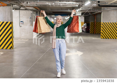 A happy woman wearing a hijab is carrying shopping bags in a parking garage, celebrating her purchases 135941838