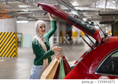 A smiling woman wearing a hijab loads shopping bags into the trunk of her car in a parking garage 135941871