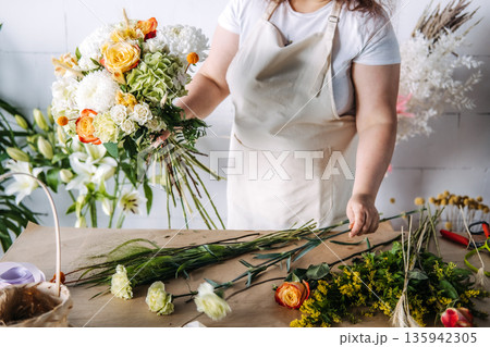 Hands of florist hold large floral bouquet over workbench in studio. Sustainable wedding decor, micro-wedding aesthetics, floral symbolism. 135942305