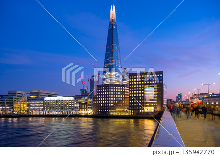 The Shard rises above the Thames River as day turns to night in London. People move along the walkway, enjoying the city as the lights begin to shine. This scene captures urban life. The Shard rises above the Thames River as day turns to night in London. People move along the walkway, enjoying the city as the lights begin to shine. This scene captures urban life. 135942770