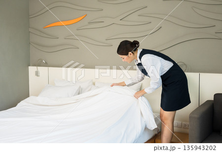 Hotel woman housekeeper in uniform neatly making bed with white linens in modern guest room Hotel woman housekeeper in uniform neatly making bed with white linens in modern guest room 135943202