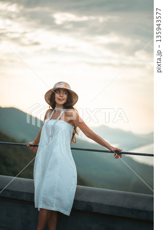 Charming woman in white dress posing at mountain sunset view, Beautiful female with straw hat against golden hour mountains background Charming woman in white dress posing at mountain sunset view, Beautiful female with straw hat against golden hour mountains background 135943577