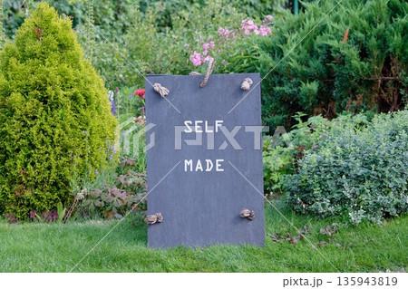 A handmade sign stands in a lush garden, its dark surface contrasting with the vibrant colors of flowers and greenery, celebrating the spirit of being self made and resourceful 135943819