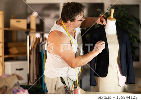 Middle-aged male dressmaker fitting jacket on mannequin Middle-aged male dressmaker fitting jacket on mannequin 135944125