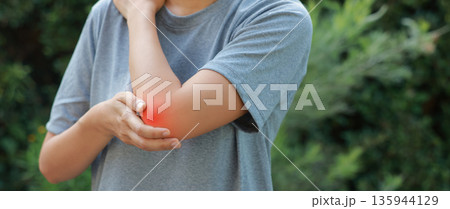 Close-up of woman hand holding painful injured elbow with red inflamed spot. Concept of joint bone pain, muscle injury, arthritis, tendonitis, or ache from sports or work in outdoor background. 135944129