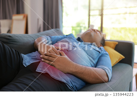 Overweight man lying on sofa holding stomach with red inflamed spot from abdominal pain. Concept of indigestion, gastritis, food poisoning, stomachache, and unhealthy lifestyle in living room. Overweight man lying on sofa holding stomach with red inflamed spot from abdominal pain. Concept of indigestion, gastritis, food poisoning, stomachache, and unhealthy lifestyle in living room. 135944223
