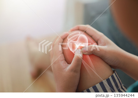 Close-up of child hand holding painful injured knee with red inflamed spot and X-ray bone skeleton joint overlay. Medical concept of pediatric fracture, arthritis, or muscle ache in indoor background. 135944244