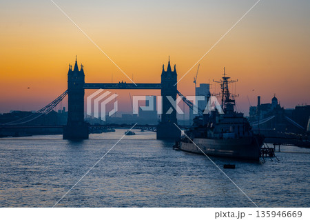 The scene shows dawn at the River Thames, featuring the silhouette of Tower Bridge and HMS Belfast. The sky has light colors as boats move on the water in London. 135946669
