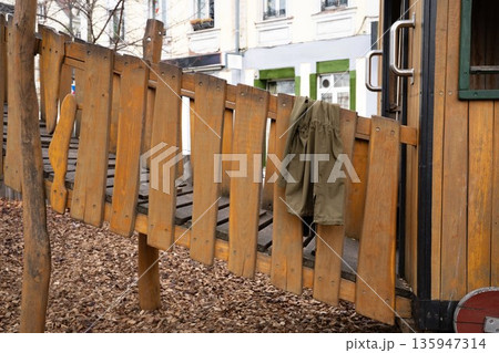 Olive parka coat hanging on a wooden children playground bridge outdoors. Everyday scene showing a personal clothing item, representing casual style, autumn, and outdoor life. Olive parka coat hanging on a wooden children playground bridge outdoors. Everyday scene showing a personal clothing item, representing casual style, autumn, and outdoor life. 135947314