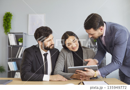 Office people, couple, colleagues gathered around desk looking attentively at tablet computer Office people, couple, colleagues gathered around desk looking attentively at tablet computer 135949772
