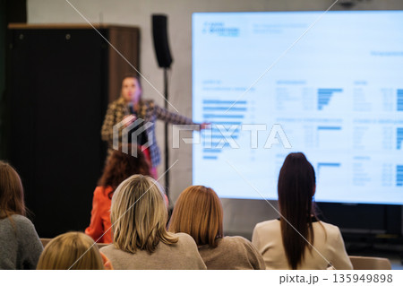 Speaker presents data to audience in a conference setting with a bright screen and attentive listeners 135949898