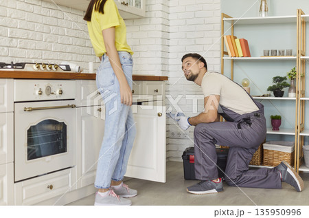 Man plumber fixing broken pipe in modern kitchen and talking with female client at home. 135950996