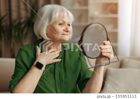 Elderly caucasian woman touching neck while looking at mirror Elderly caucasian woman touching neck while looking at mirror 135951113